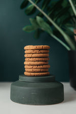 Load image into Gallery viewer, Stack of ginger biscuits displayed on a green pedestal with a dark green background and plant decor.
