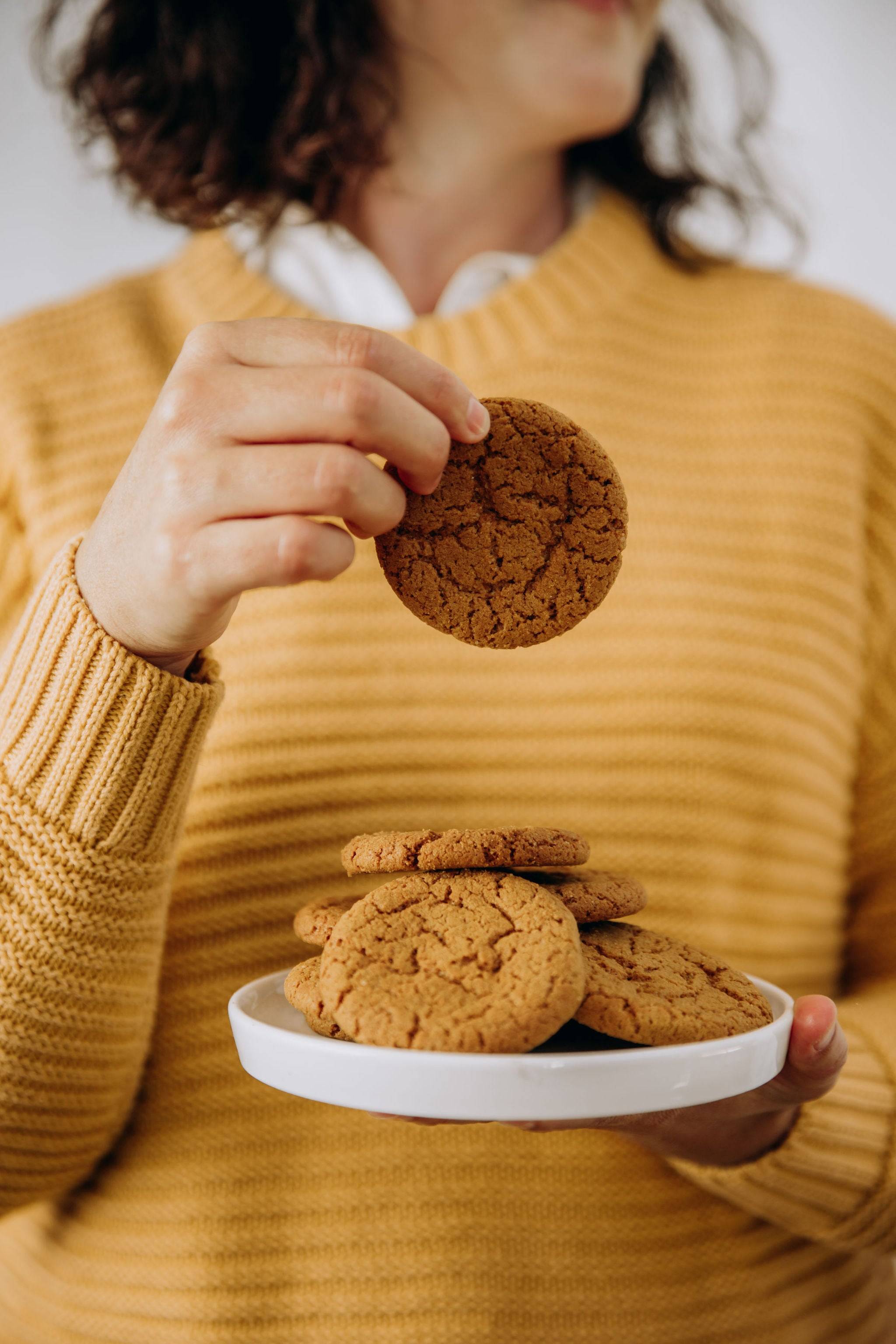 Person in a mustard sweater holding a plate of ginger biscuits, lifting one cookie to enjoy.